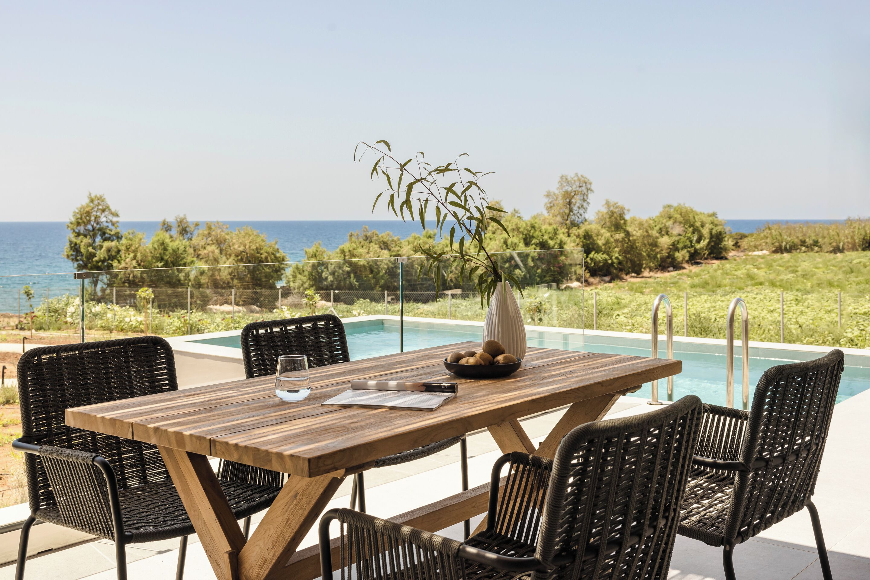A wooden table with chairs and a vase of flowers, overlooking a swimming pool with a view of the sea and greenery.