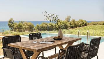 Mesa de madeira com cadeiras e um vaso de flores, com vista para uma piscina e o mar, além de vegetação.