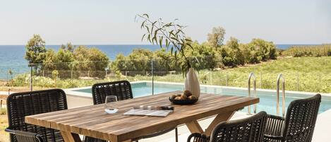 A wooden table with chairs and a vase of flowers, overlooking a swimming pool with a view of the sea and greenery.