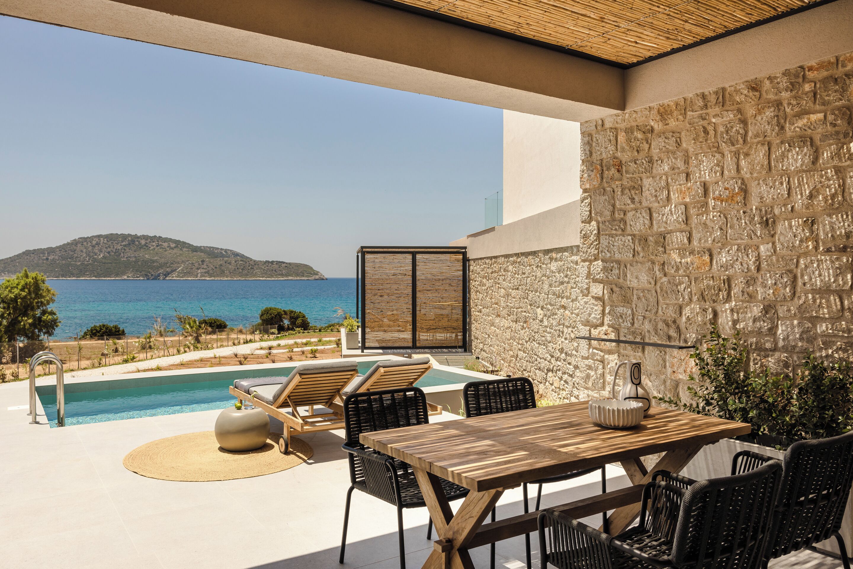 Un espace aménagé au bord de la piscine, avec une table et des chaises en bois, un mur en pierre et une vue sur la mer.