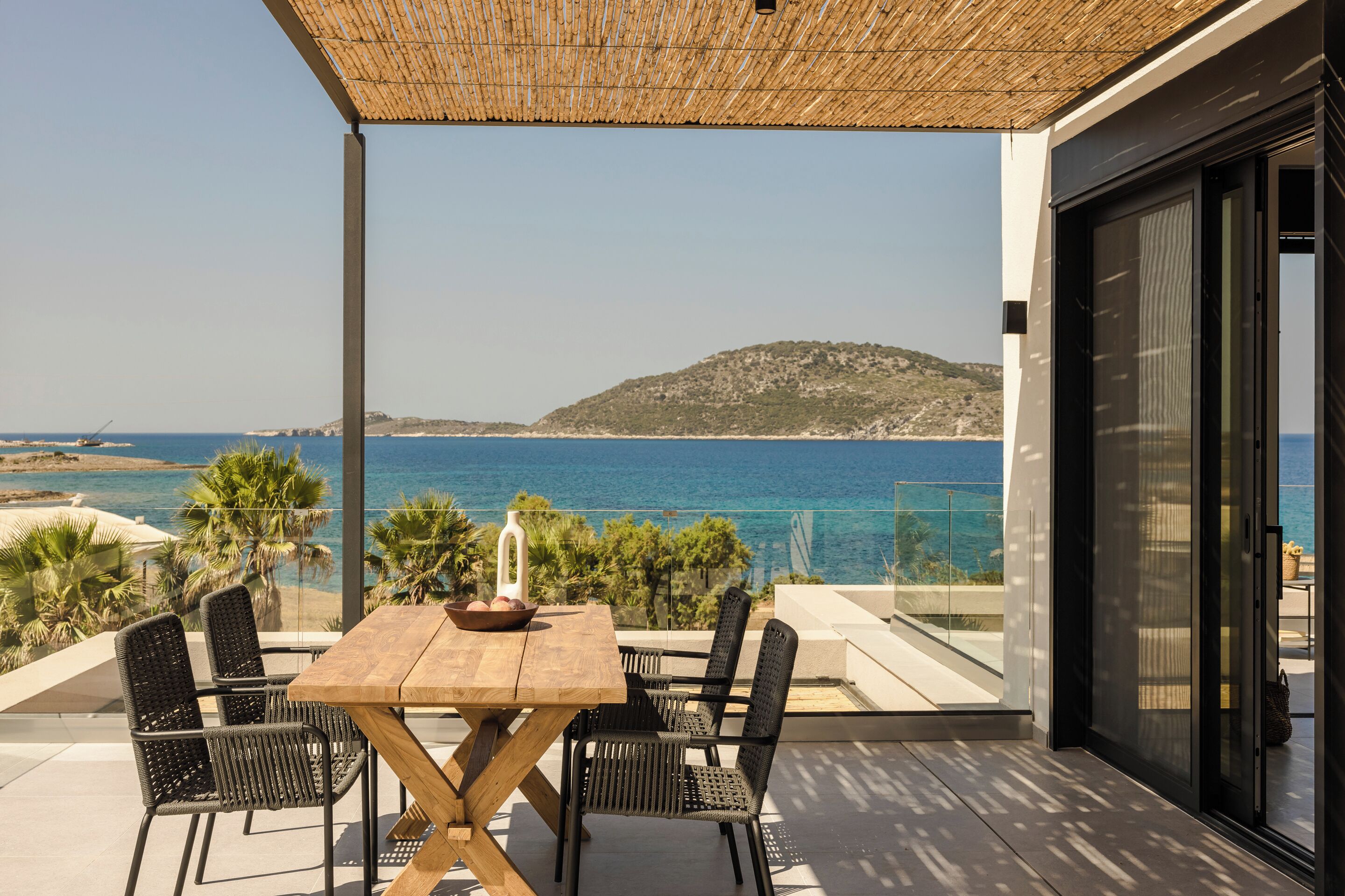 Una terraza con mesa y sillas de madera, con vista al mar y una colina distante.