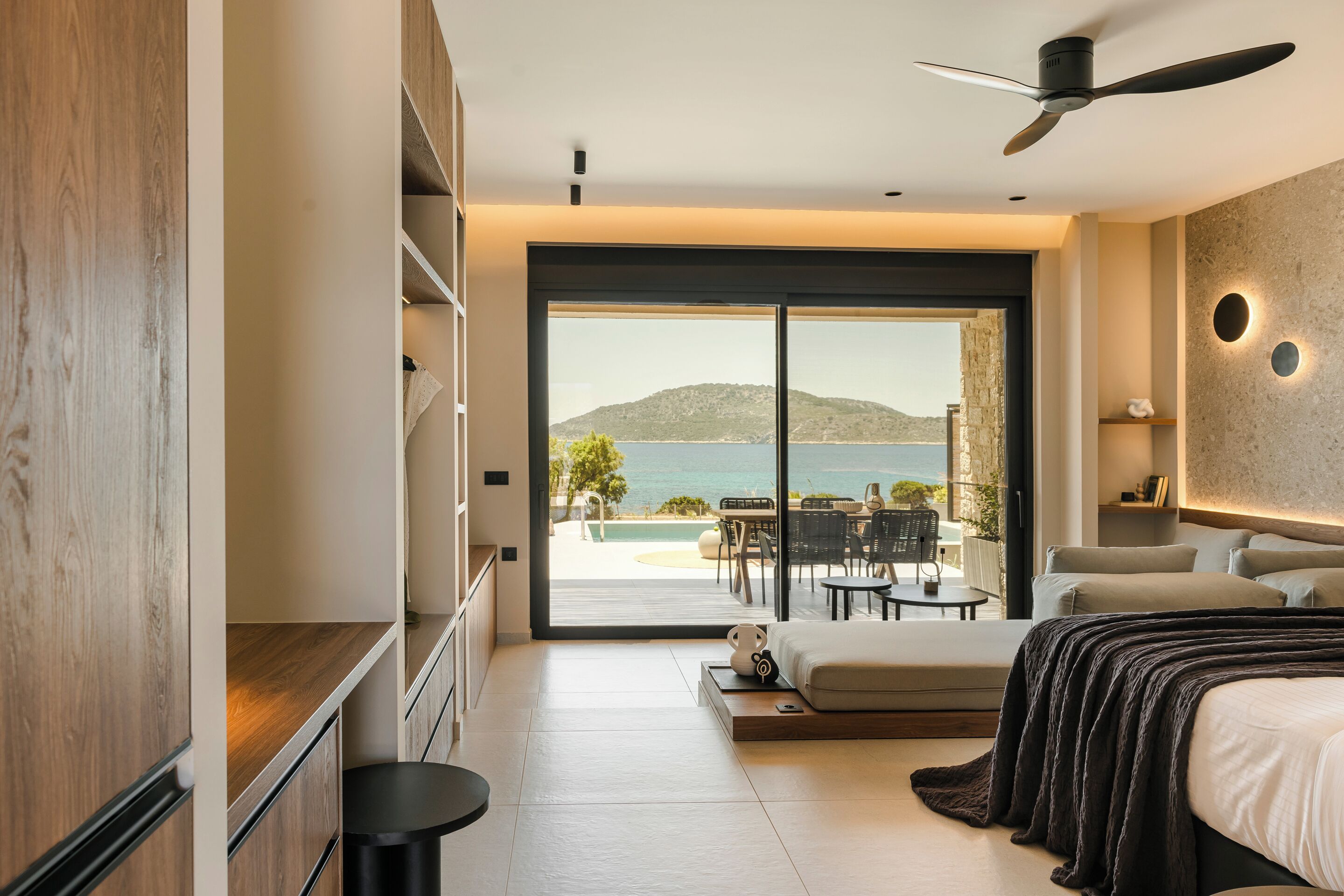 A modern bedroom with a large sliding glass door leading to a balcony with a pool and a view of the sea.