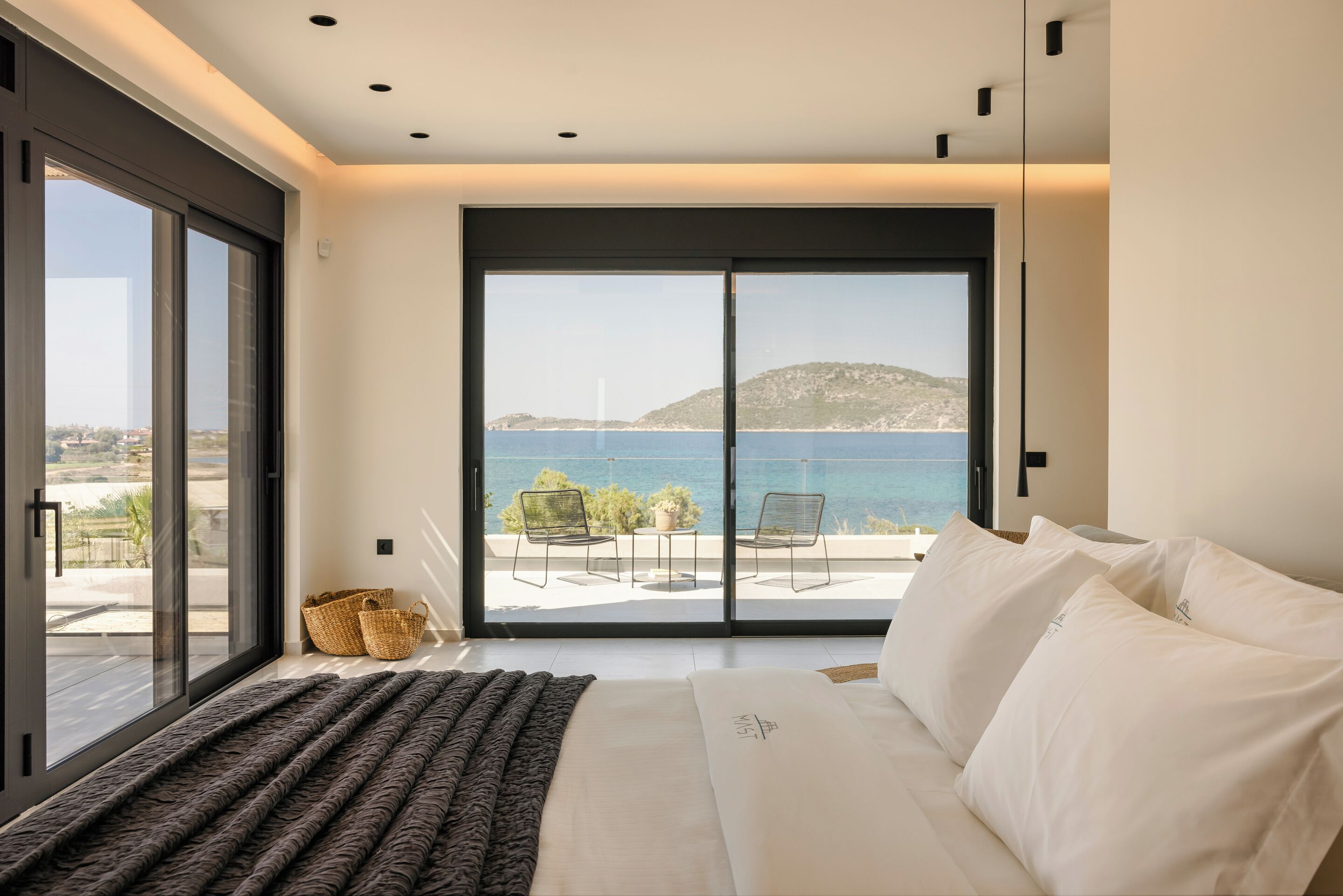 A bedroom with a large sliding glass door leading to a balcony with a view of the sea and a distant hill.