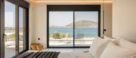 A bedroom with a large sliding glass door leading to a balcony with a view of the sea and a distant hill.