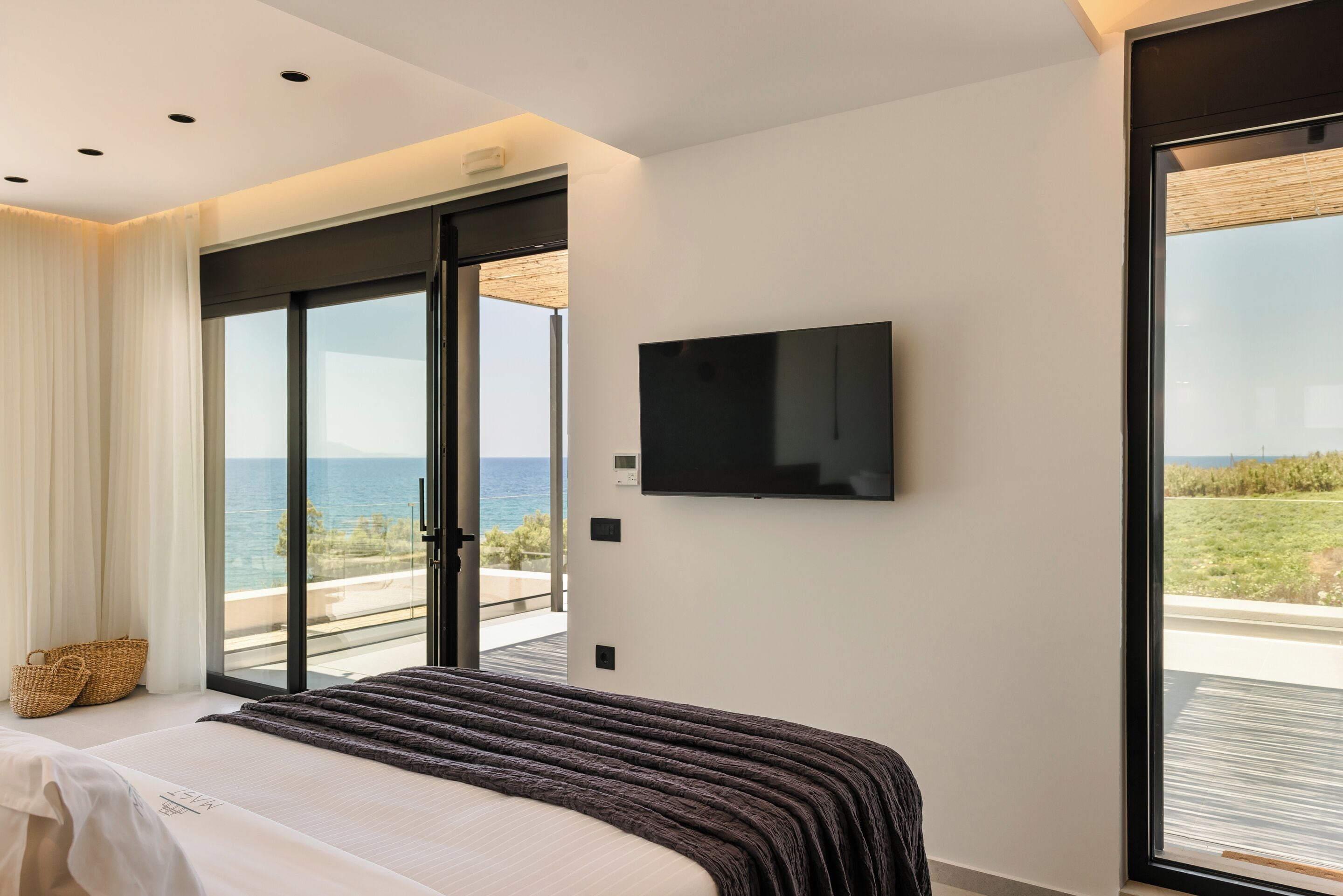 A modern bedroom with a large bed, a flat-screen TV mounted on the wall, and a sliding glass door leading to a balcony with a view of the sea.