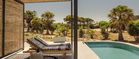 Un área junto a la piscina con sillones de descanso y una pequeña mesa, rodeada por una cerca de bambú y palmeras.