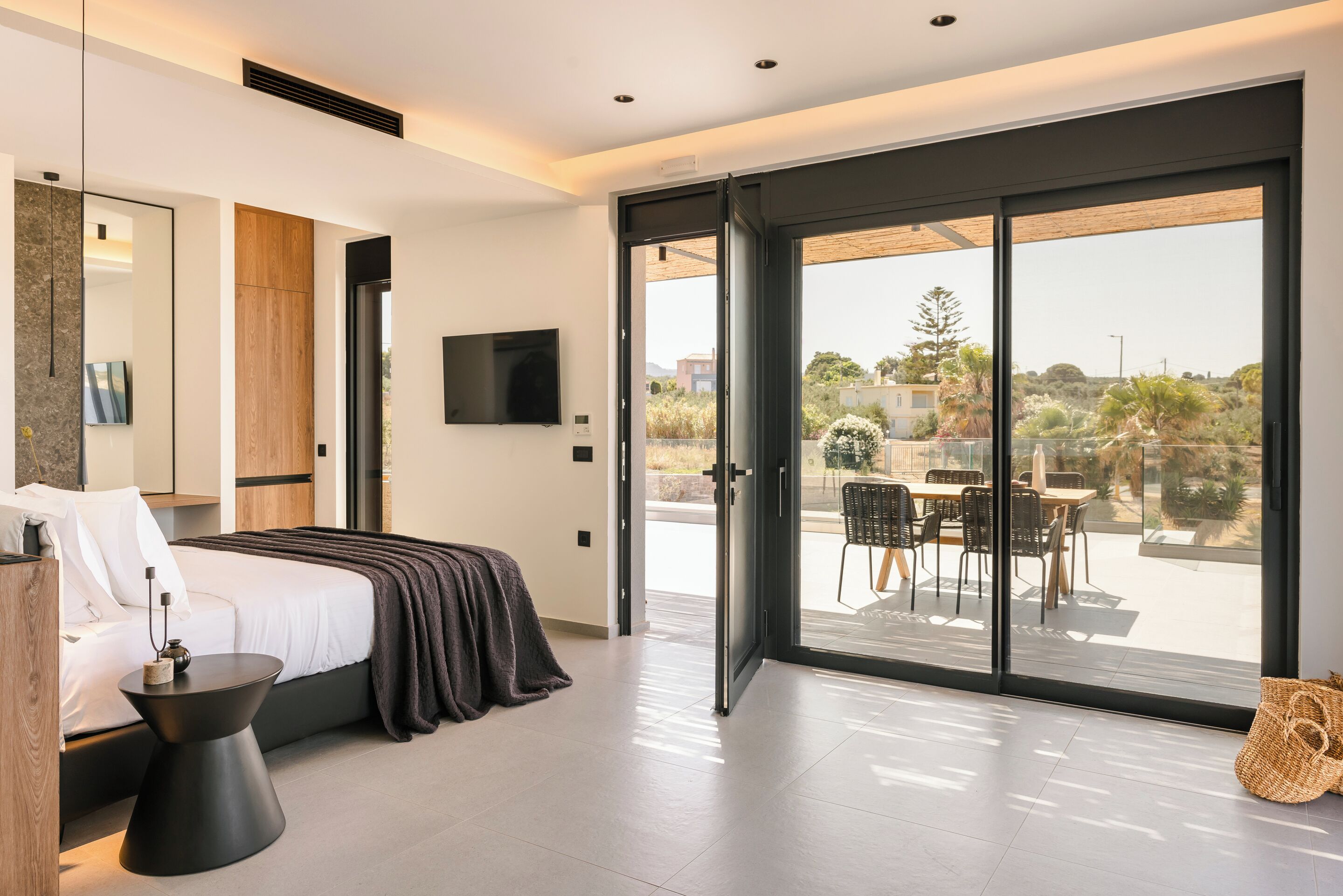Un dormitorio moderno con cama, mesita de noche y un amplio ventanal corredera que da a un patio exterior con mesa y sillas.