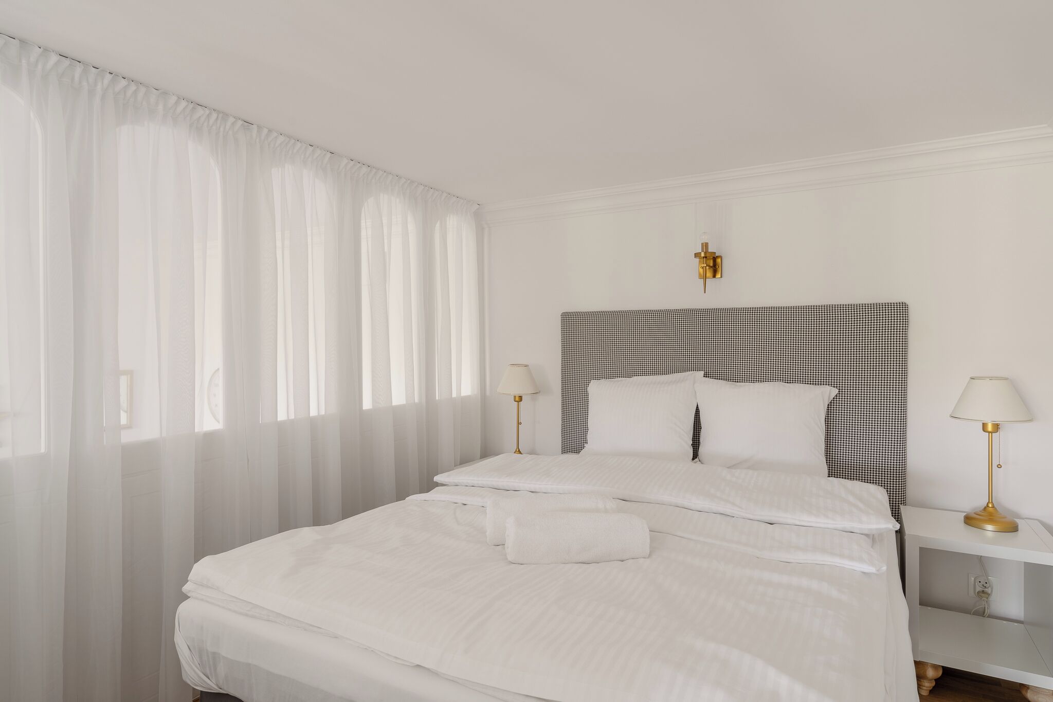A neatly made bed with white linens and two pillows, a bedside table with a lamp, and sheer curtains on the window.