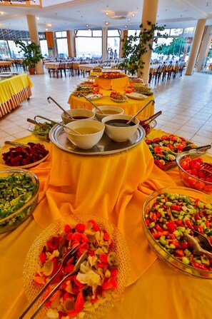 Ein reichhaltiges Buffet mit verschiedenen Speisen und Schalen auf einer gelben Tischdecke in einem großzügigen Raum mit großen Fenstern und Stühlen.