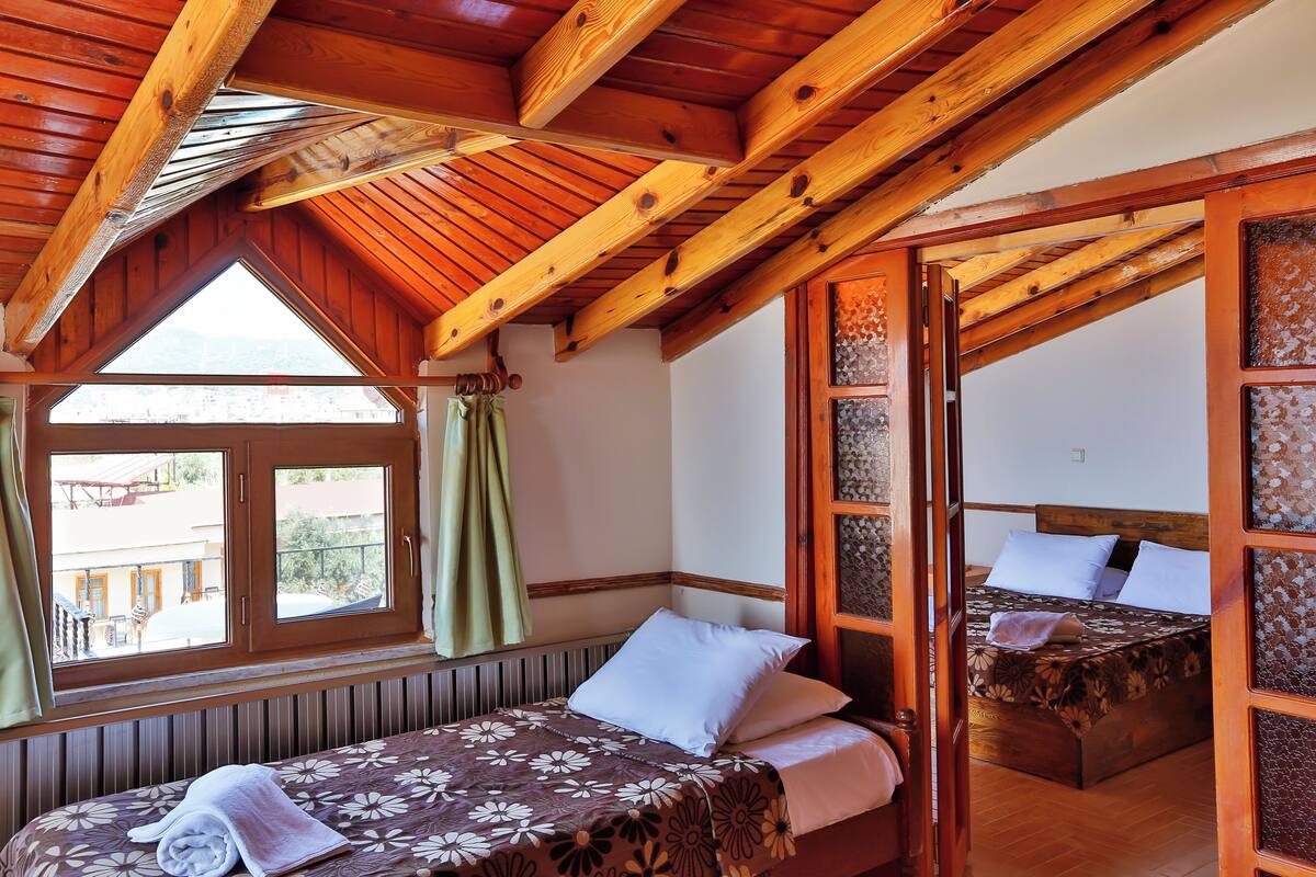 A room with a wooden ceiling, a bed with a floral bedspread, a window with curtains, and a view of a building outside.
