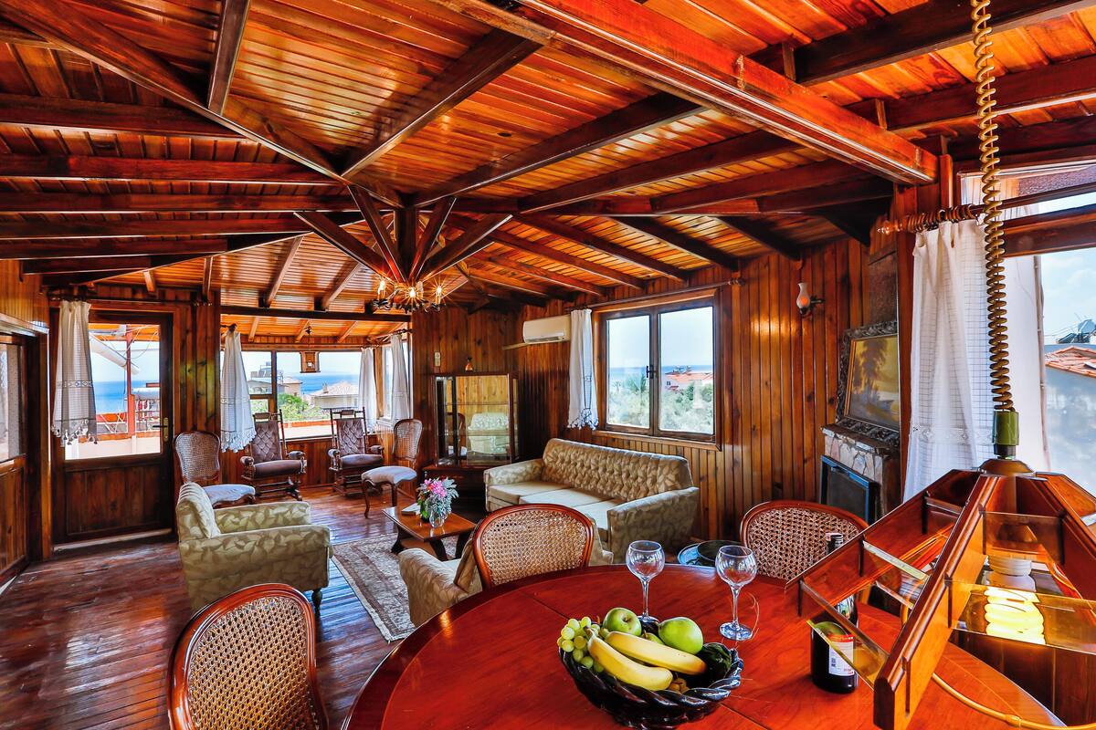 A wooden interior with a round dining table, wicker chairs, a sofa, and a large window overlooking the sea.