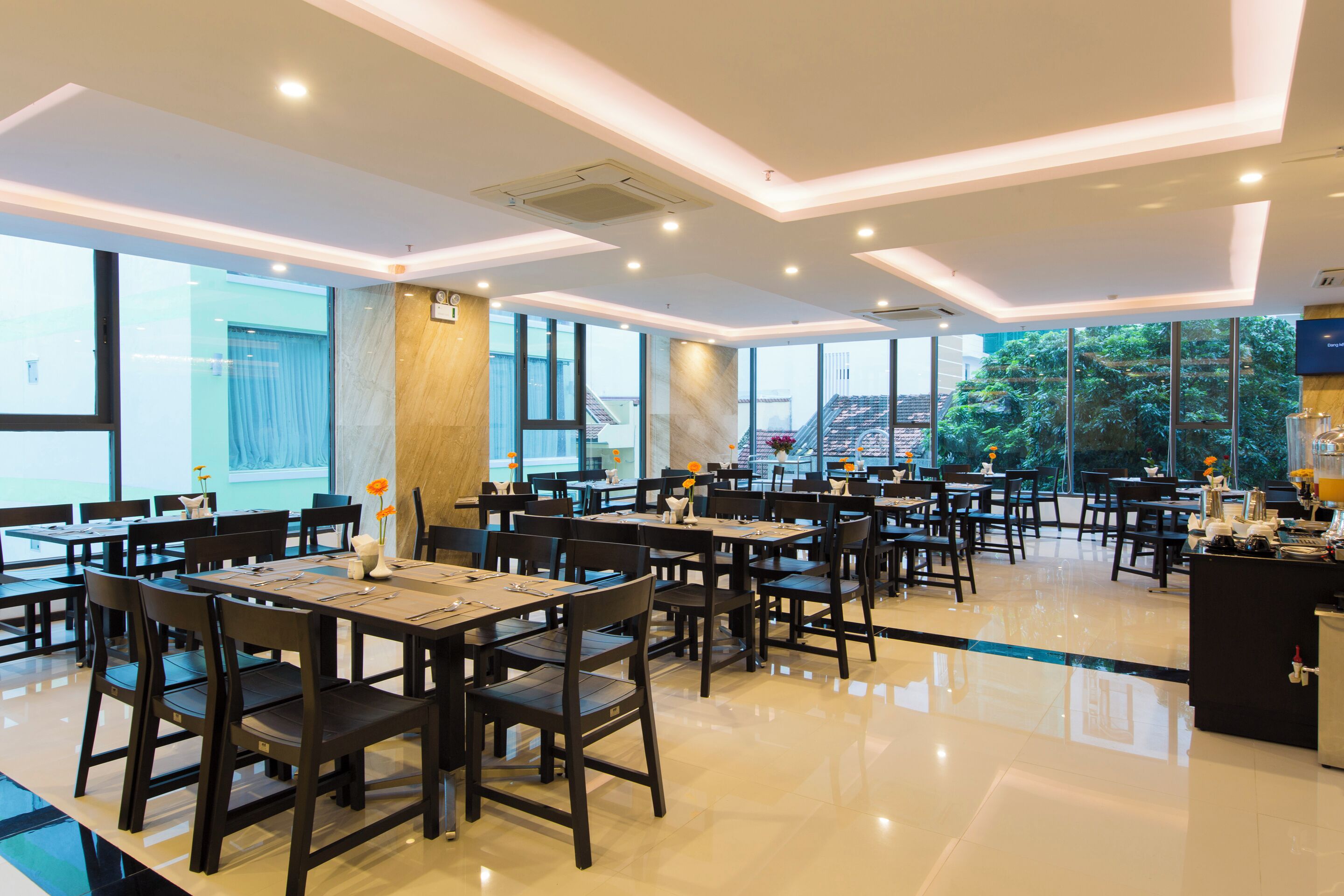 A spacious dining area with multiple tables and chairs, large windows offering views of the outside, and a modern ceiling design.