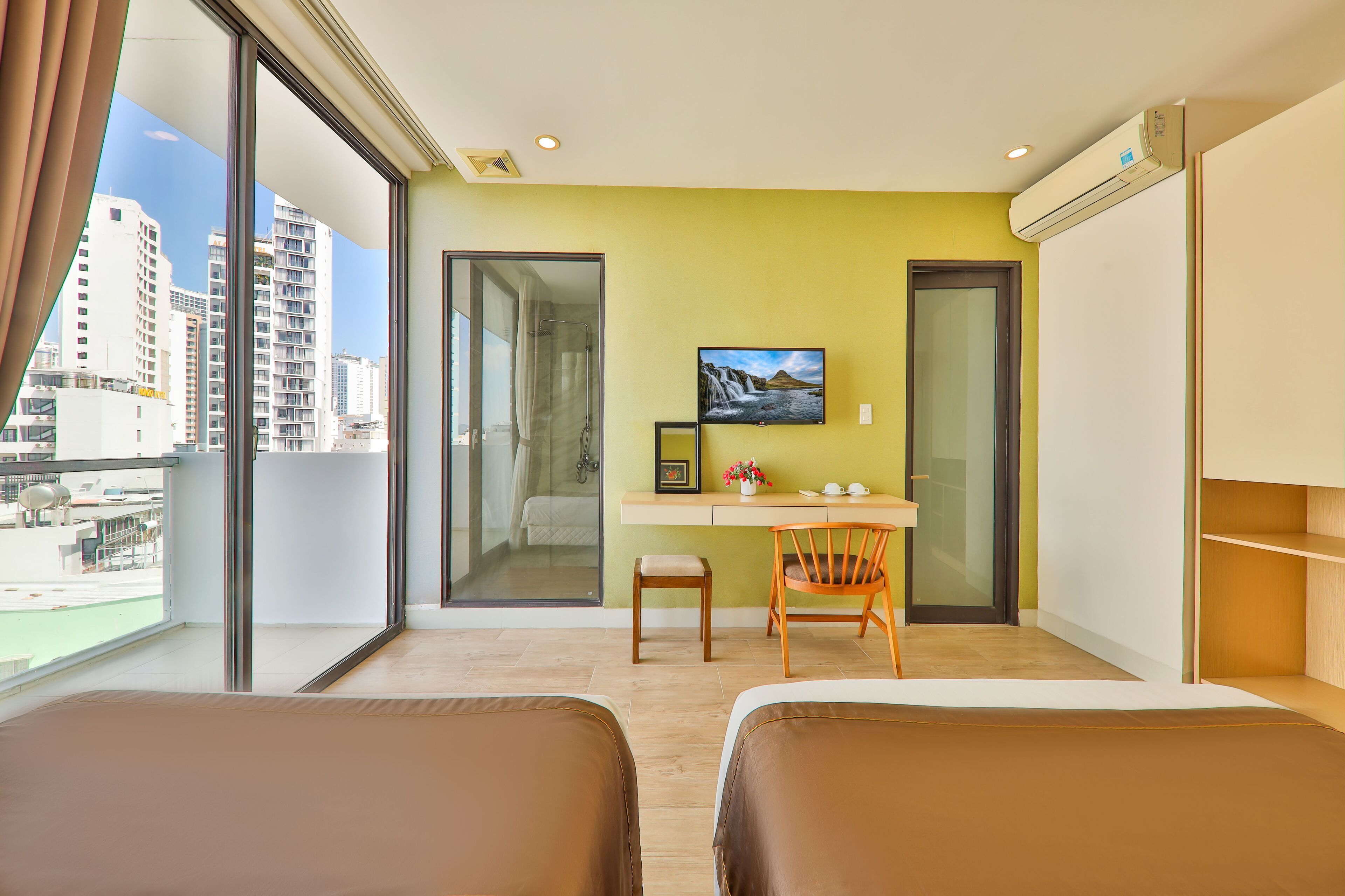 A hotel room with two beds, a desk with a chair, a TV, and a large window with a city view.