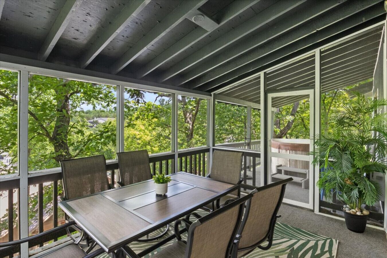 A covered outdoor patio with a table and chairs, surrounded by glass walls and a view of trees.