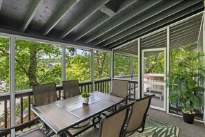 A covered outdoor patio with a table and chairs, surrounded by glass walls and a view of trees.