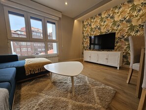 A living room with a sofa, a coffee table, a TV on a white cabinet, and a large window with a view of buildings. - 1A126 ALQ TEMPORADA  centrico en Gijon (Gijón)