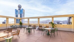 Une terrasse sur le toit avec des tables et des chaises, de grandes fenêtres offrant une vue sur la ville et un bâtiment moderne en arrière-plan.