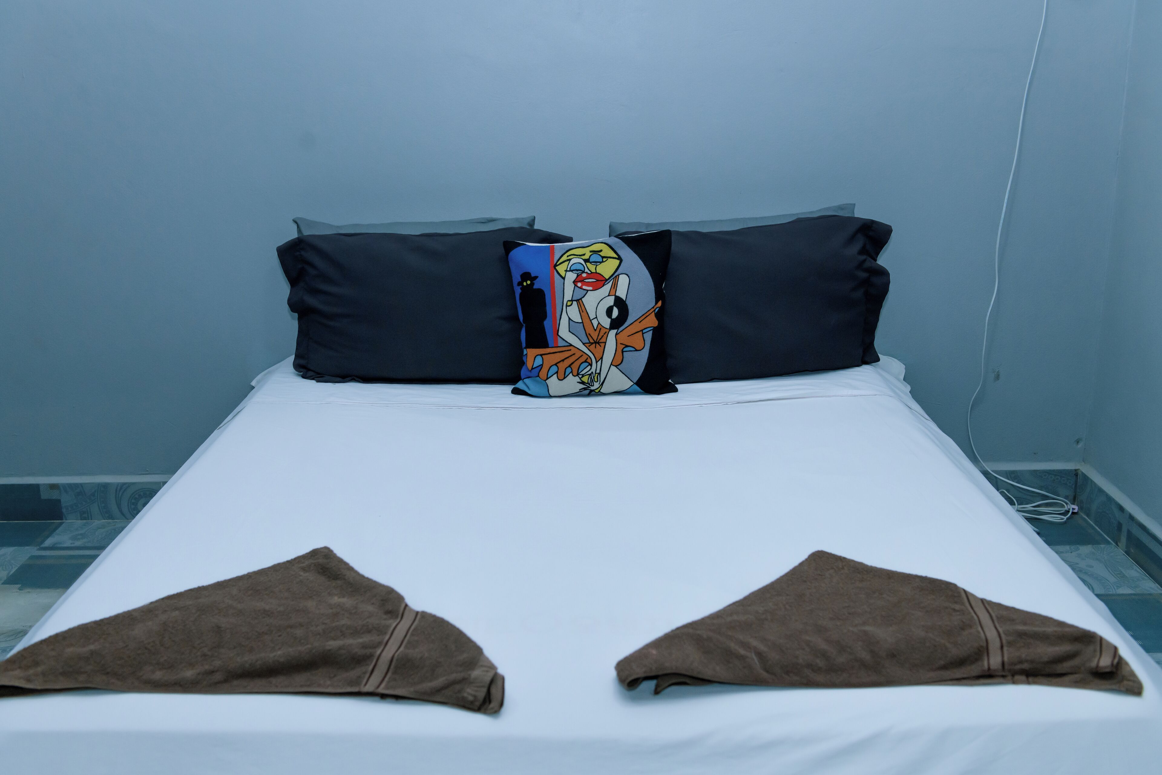 A bed with a white sheet, two dark pillows, and a colorful pillow with a cartoon character. There are brown towels at the foot of the bed.