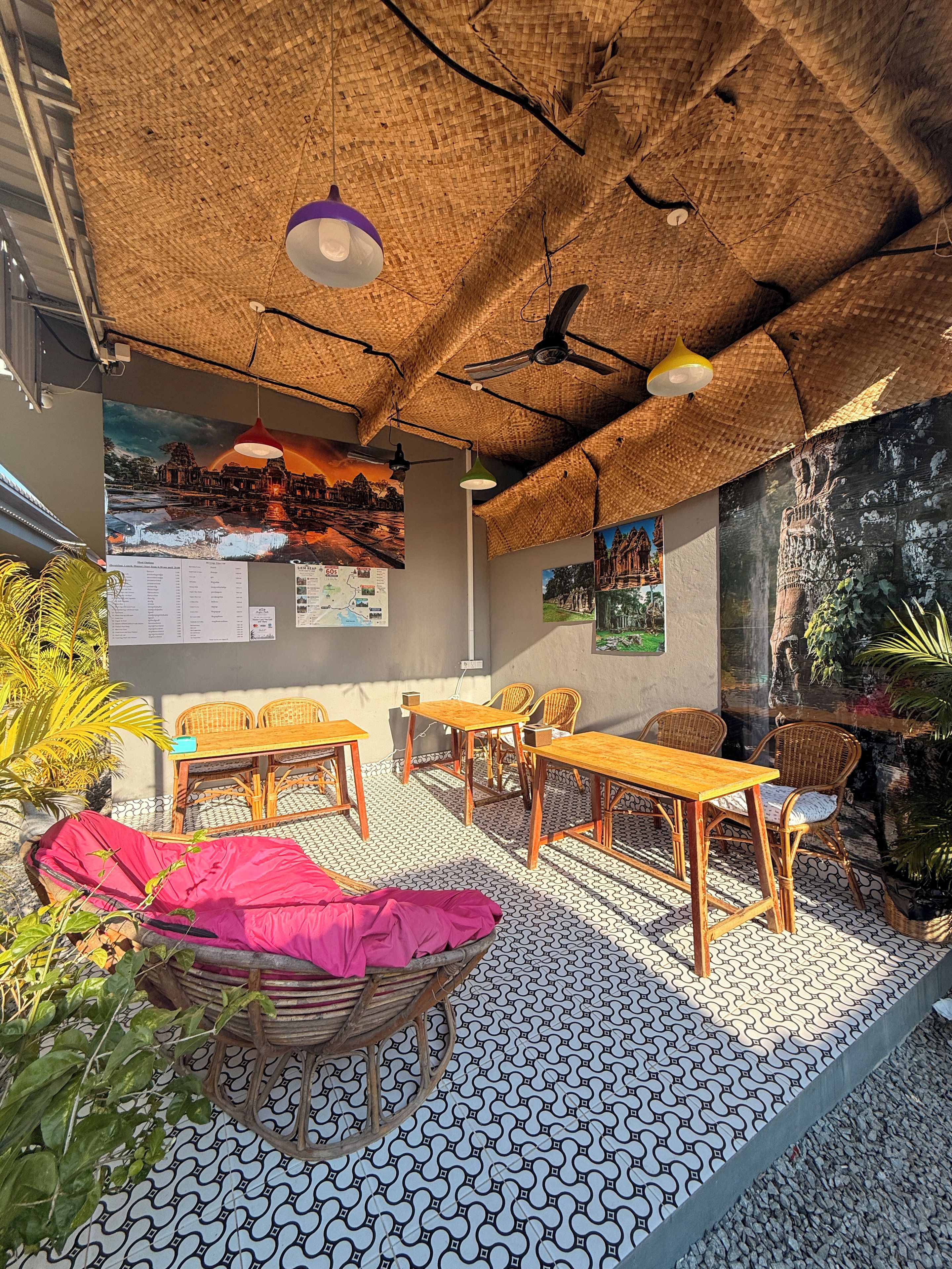 Un espacio al aire libre con mesas y sillas, techo de paja y una pared con folletos informativos.