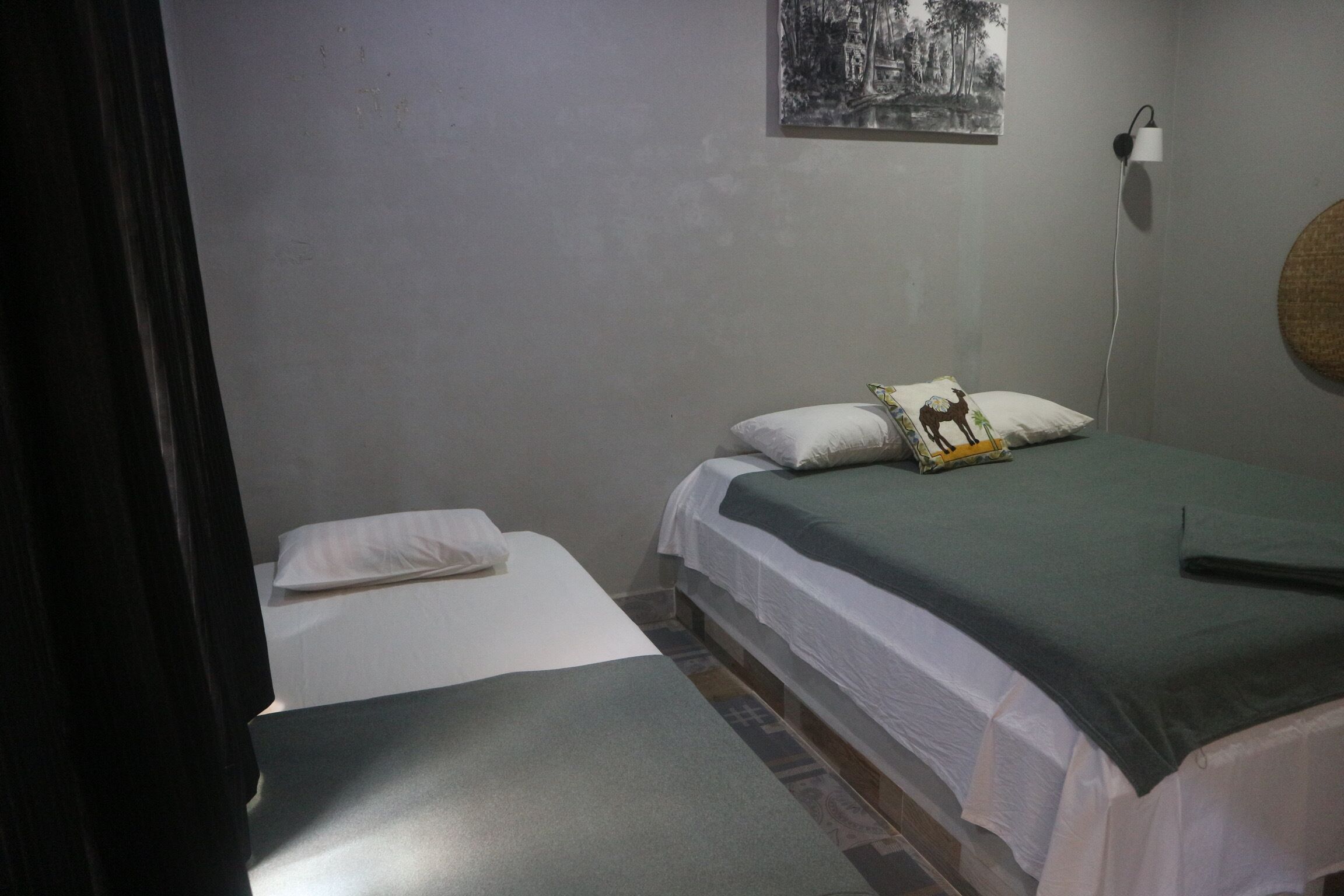 Une chambre avec deux lits, l’un recouvert d’une couvre-lit verte et l’autre d’une couvre-lit blanche. Il y a une lampe fixée au mur et un tableau au-dessus des lits.