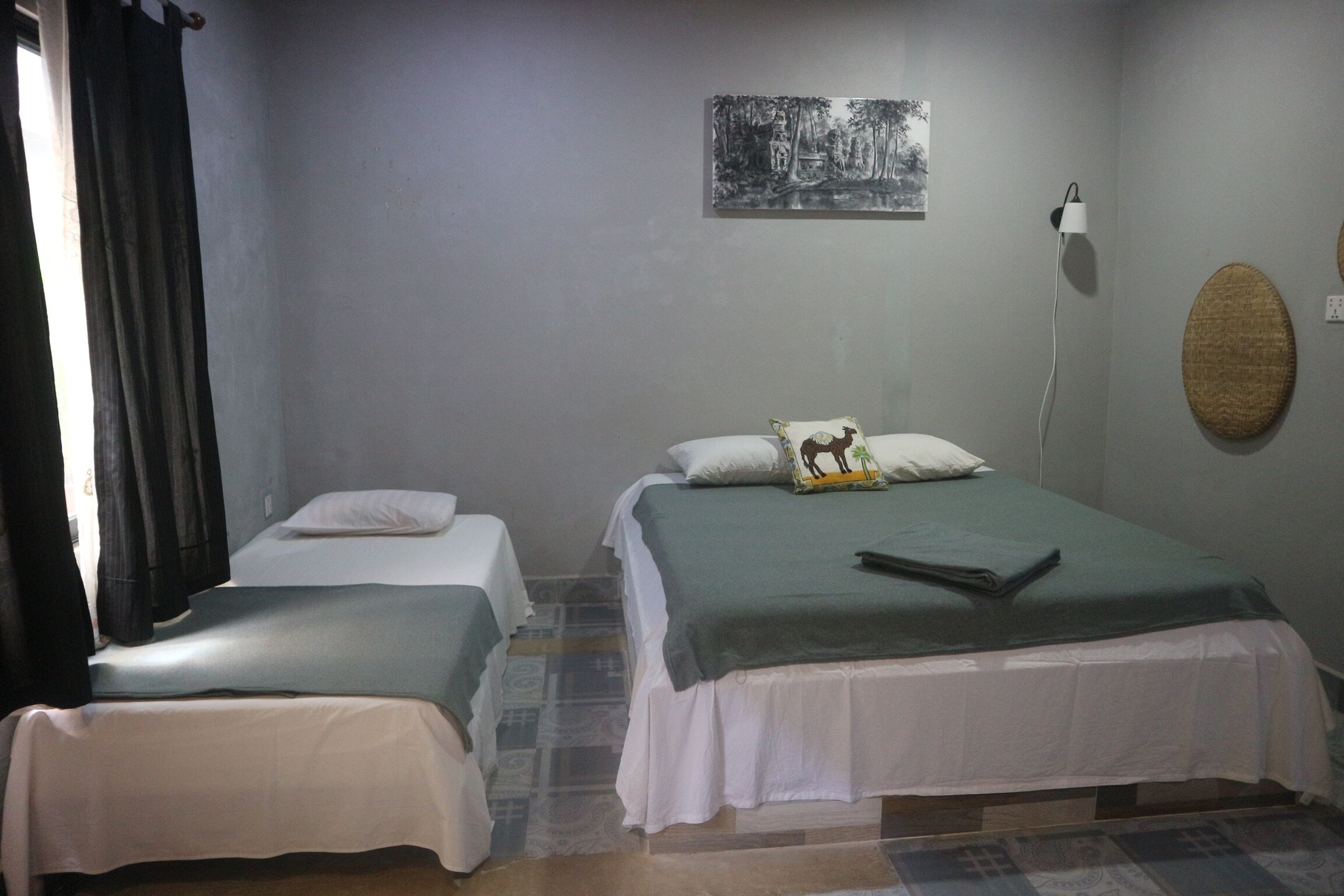 A room with two beds, a wall-mounted lamp, a painting, and a woven wall decoration.