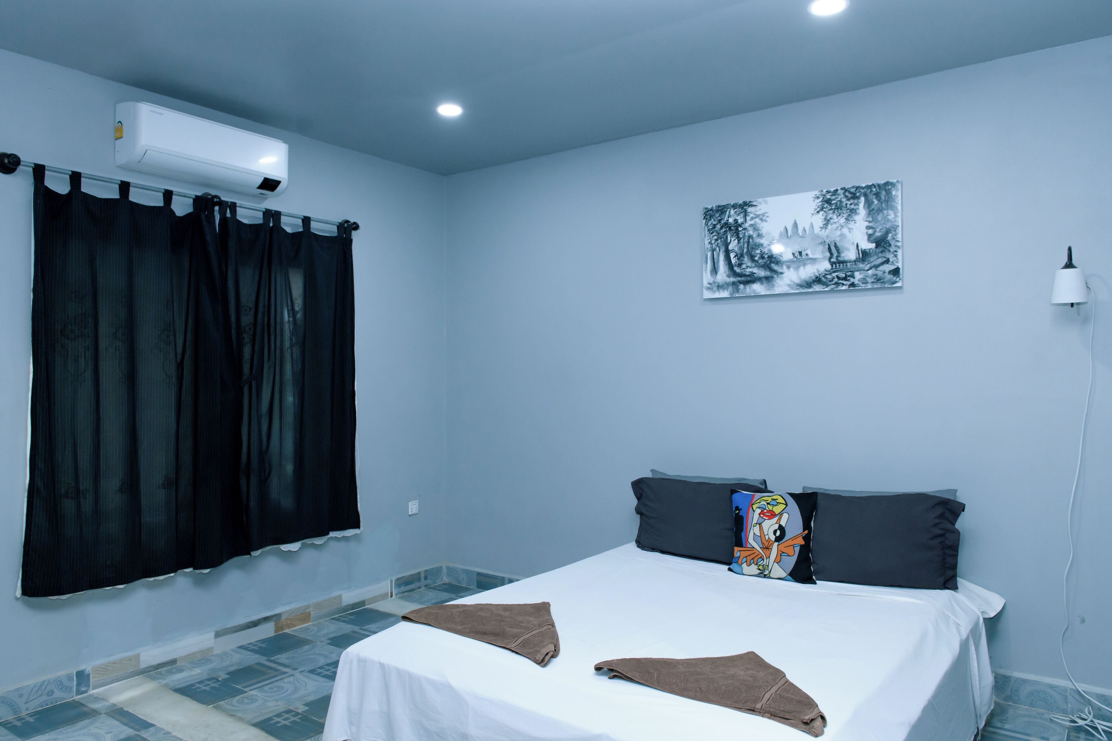 A bedroom with a bed, black curtains, a wall-mounted air conditioner, a framed picture, and a bedside lamp.