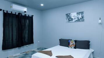 A bedroom with a bed, black curtains, a wall-mounted air conditioner, a framed picture, and a bedside lamp.