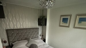 Une chambre à coucher avec une tête de lit capitonnée, un grand lit, un lustre, des tableaux encadrés au mur et un petit tabouret.