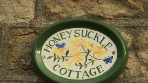 Eine runde, grüne Plakette mit floralen und Schmetterlingsmotiven sowie dem Text "Honey Suckle Cottage" an einer Ziegelwand.