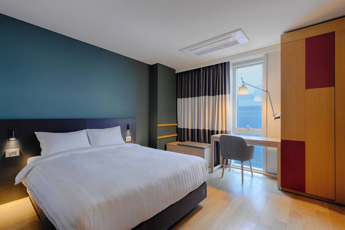 A modern hotel room with a large bed, bedside table, desk, chair, and a wardrobe.