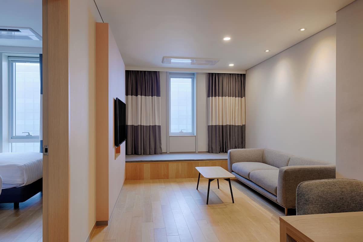 A modern hotel room with a bed, a sofa, a small table, and a TV mounted on the wall.