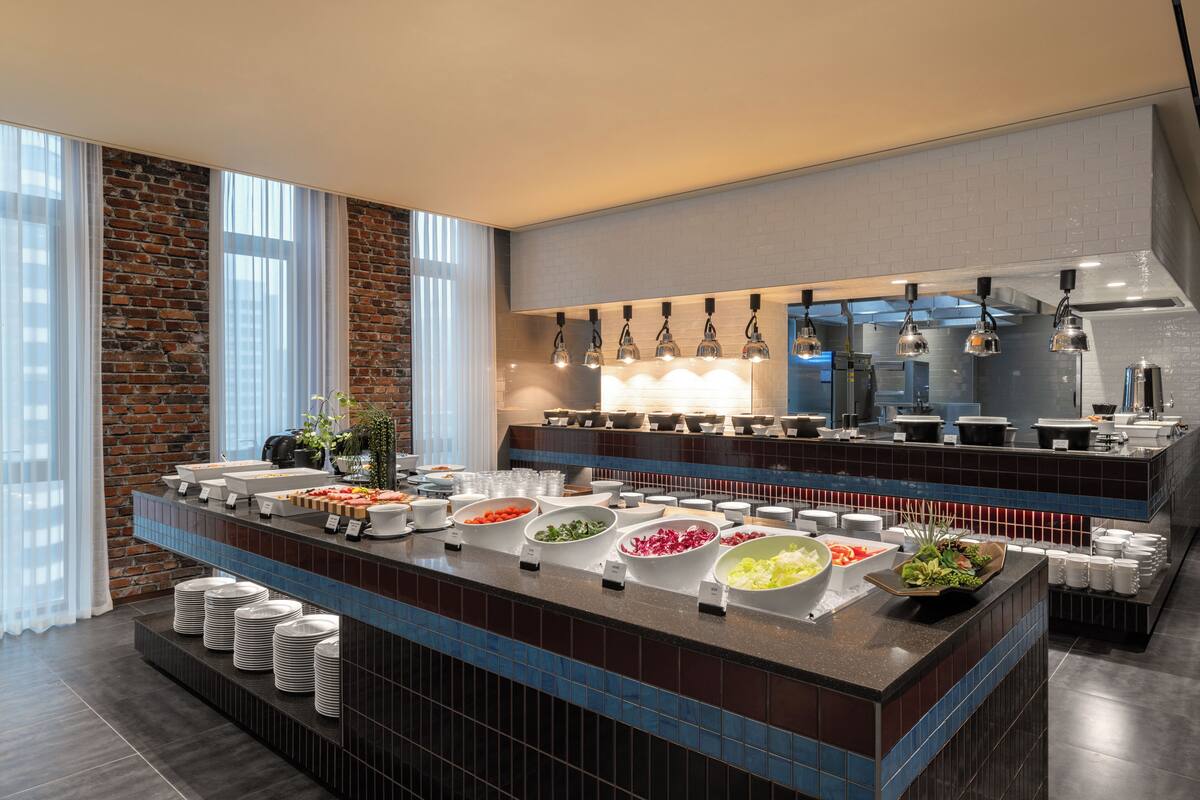 A modern buffet setup with various food stations, a brick wall, and large windows.