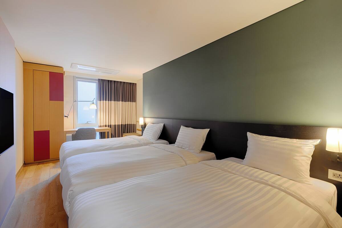 A hotel room with three beds, a desk, a chair, and a window with curtains.