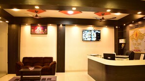 A reception area with a dark reception desk, a brown sofa, a small table with a bowl, a flat-screen TV, and ceiling fans. - HOTEL JYOTI PALACE (Biaora)