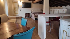 Una habitación acogedora con una mesa de comedor de madera, sillas azules, una cama con estructura de metal, un televisor montado en la pared y una zona de cocina con armarios.