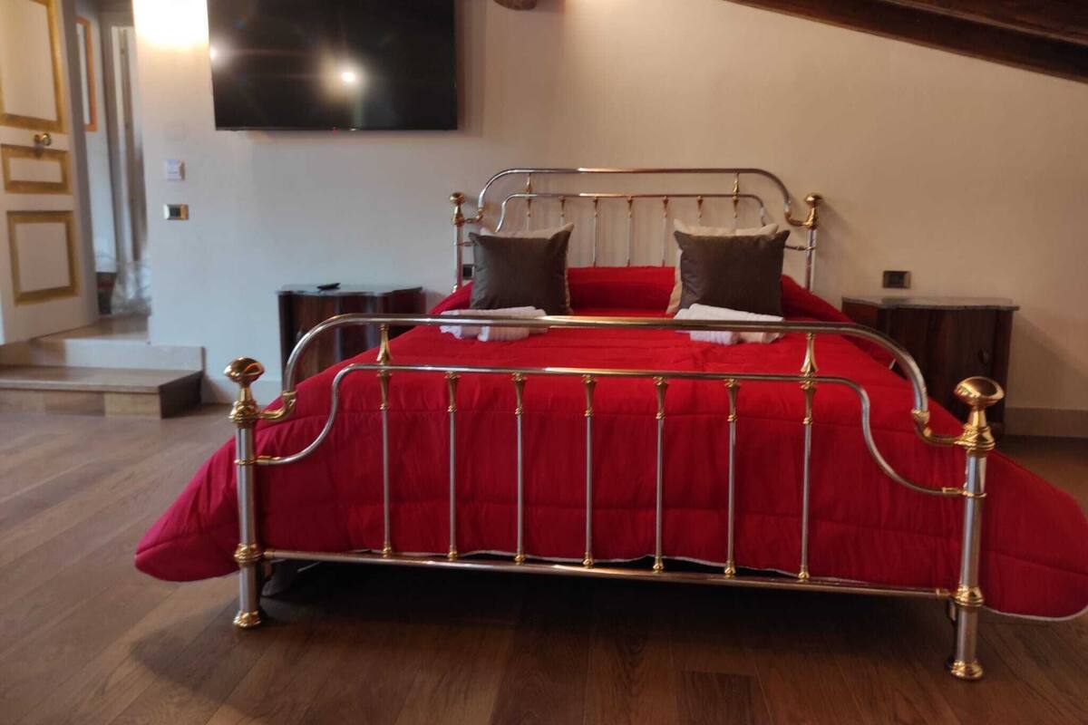A bedroom with a metal bed frame, red bedding, a flat-screen TV on the wall, and a wooden staircase in the background.