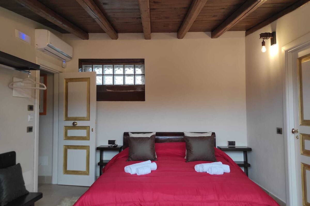 A bedroom with a wooden ceiling, a large bed with red bedding and two pillows, a nightstand, and a window with blinds.