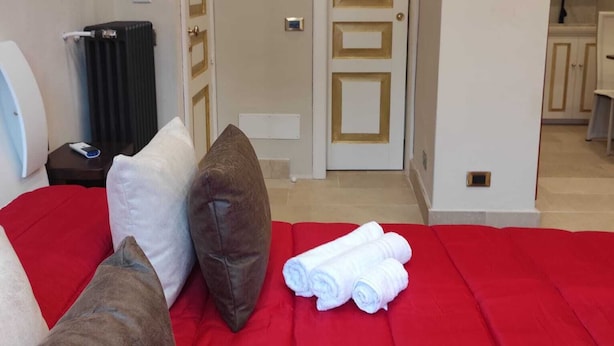 Habitación de hotel con una colcha roja, dos almohadas, un radiador, una mesita con un teléfono y vistas a una zona de cocina.