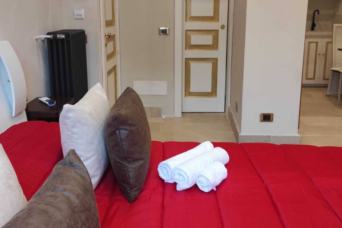 A hotel room with a red bedspread, two pillows, a radiator, a small table with a phone, and a view of a kitchen area.