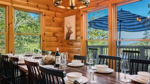 A wooden dining table set with white plates, bowls, and wine glasses, with a deer head painting on the wall and a large umbrella outside. - Spectacular Log Cabin Mtn Views Hot Tub Pool Table Theater Room Spa/Sauna (McGaheysville)