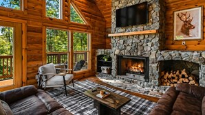 A log cabin interior with a stone fireplace, a television mounted on the wall, and a large window overlooking a forest. - Spectacular Log Cabin Mtn Views Hot Tub Pool Table Theater Room Spa/Sauna (McGaheysville)
