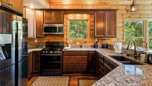 A modern kitchen with wooden cabinets, stainless steel appliances, granite countertops, and a large window providing a view of the outdoors. - Spectacular Log Cabin Mtn Views Hot Tub Pool Table Theater Room Spa/Sauna (McGaheysville)