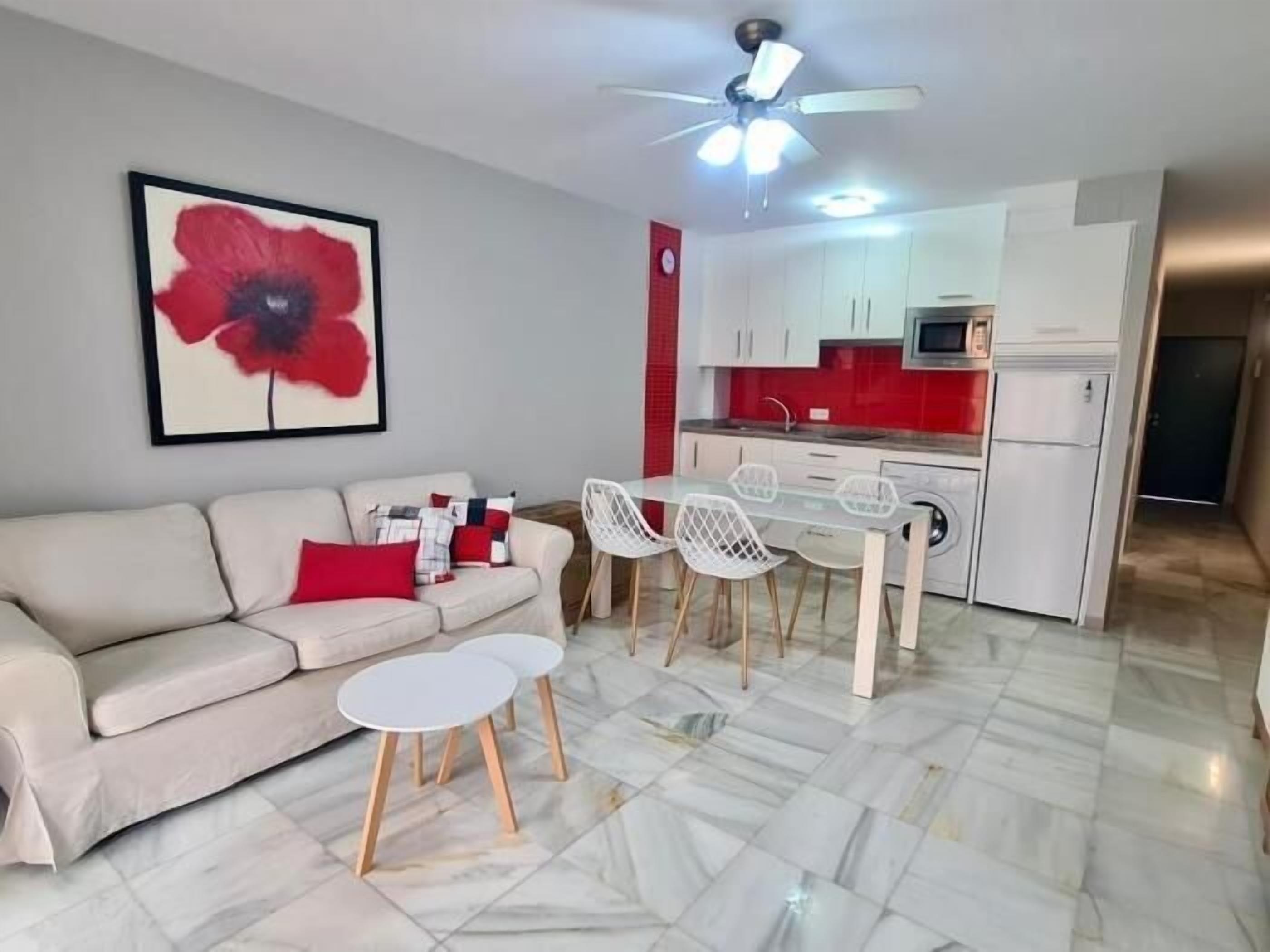A modern living room with a sofa, a dining table with chairs, a kitchen area with white cabinets, and a ceiling fan.