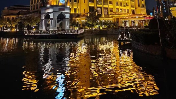 Un hotel con una torre destacada que tiene señalización iluminada, un edificio grande con varios balcones y un reflejo en el agua de enfrente.