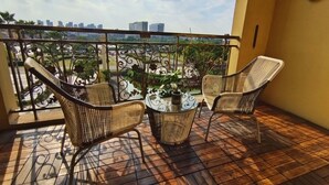 Un balcón con sillas de mimbre y una mesa de vidrio, con vistas a un paisaje urbano con edificios y zonas verdes.