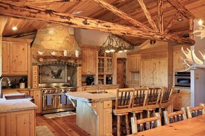 A rustic kitchen with wooden cabinets, a stone fireplace, and a large antler chandelier.