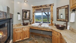 Une salle de bain avec un sol en pierre, des meubles en bois, une grande fenêtre avec vue et deux vasques surmontées de bols en bois.