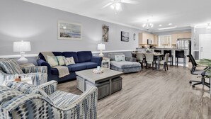 A modern living room with a blue sofa, patterned armchairs, a coffee table, and a dining area with chairs and a kitchen in the background. - Magnolia Pointe (Myrtle Beach)