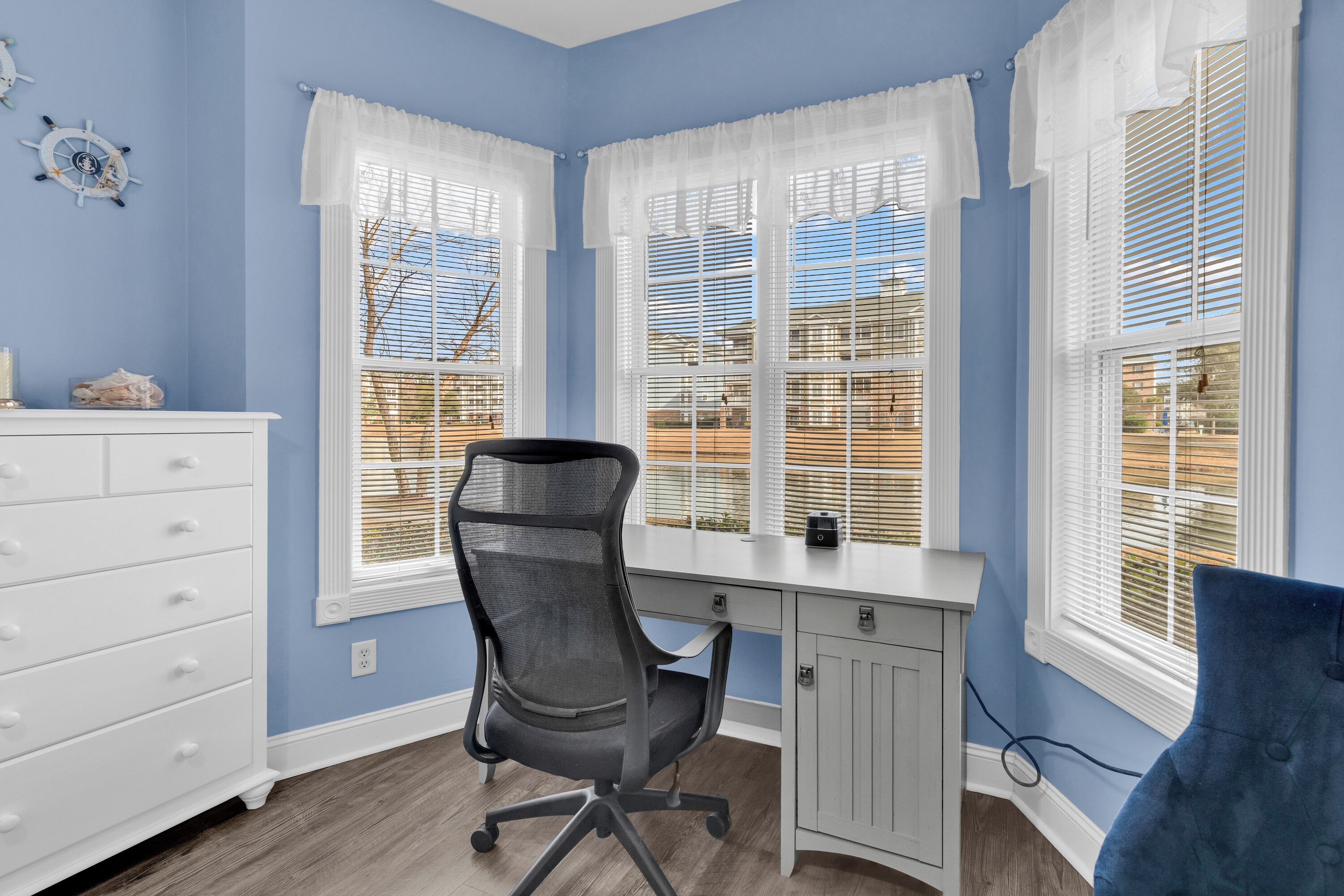 Quarto com escrivaninha, cadeira, cômoda e amplas janelas com cortinas brancas.