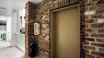 A brick wall with a beige elevator door, a white trash bin, and a small sign with an 'X' symbol.
